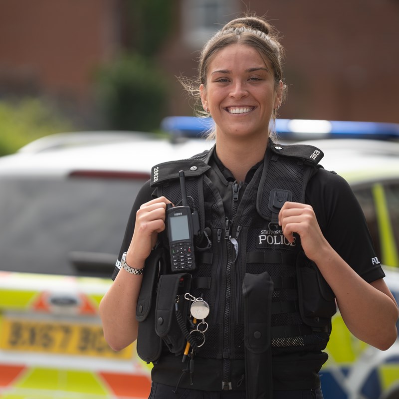 Smiling female police officer looking into the camera and stood in front of a police car.